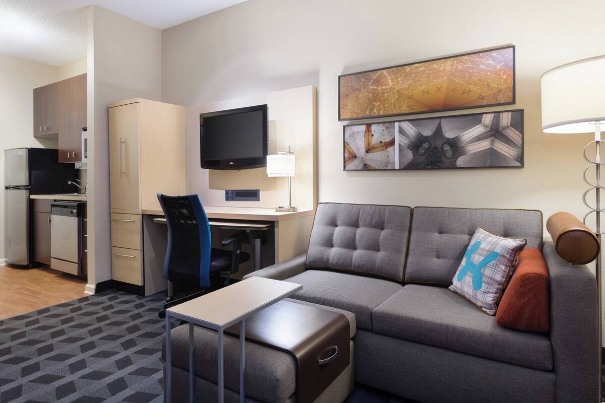 A compact hotel room with a sofa, a small desk, a television, and a kitchenette.