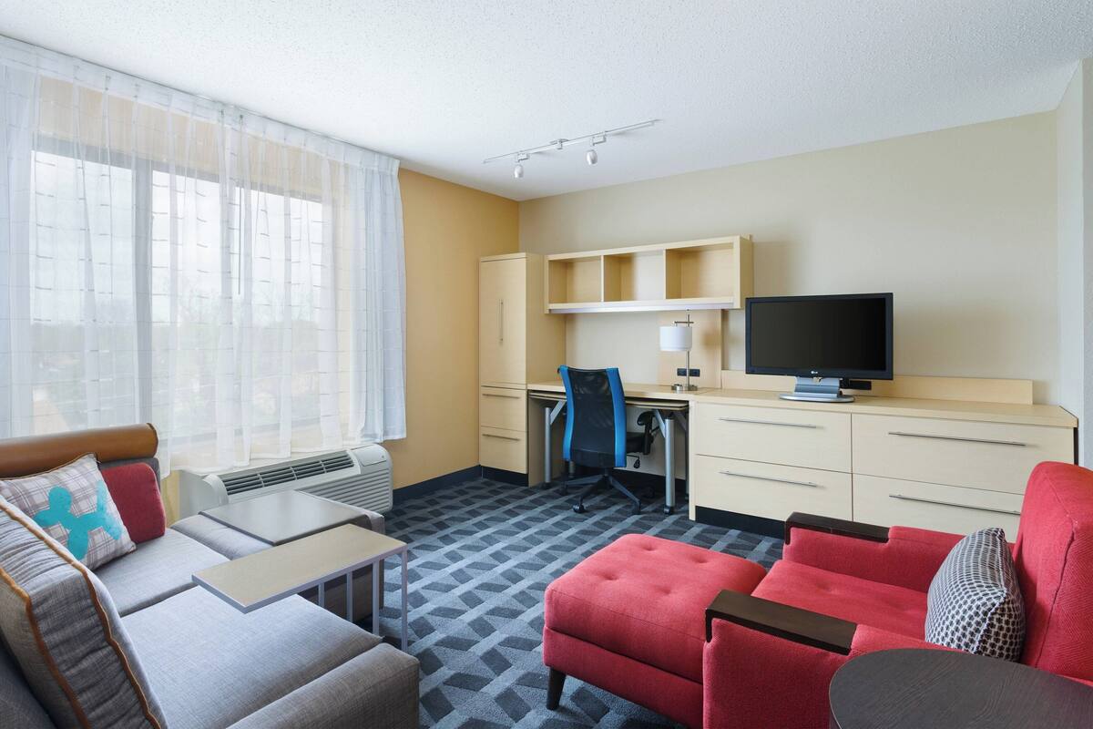 A hotel room with a sofa, armchair, and ottoman, a desk with a chair, a television, and a minibar.
