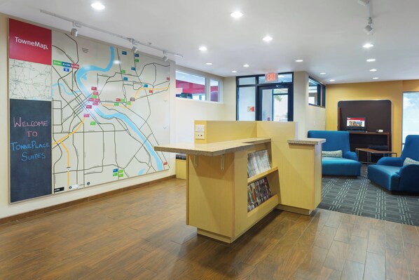 Zona de recepción de un hotel con mapa, mostrador de recepción, área de estar y pantalla de televisión.