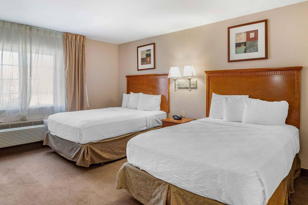 Ett hotellrum med två sängar, ett fönster med tunna gardiner, en vägglampa och en inramad tavla.