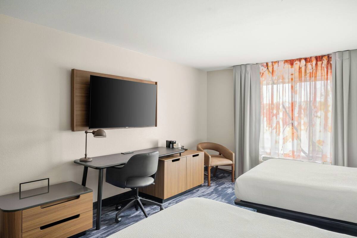 A hotel room with a bed, a desk with a chair, a television, and a window with curtains.