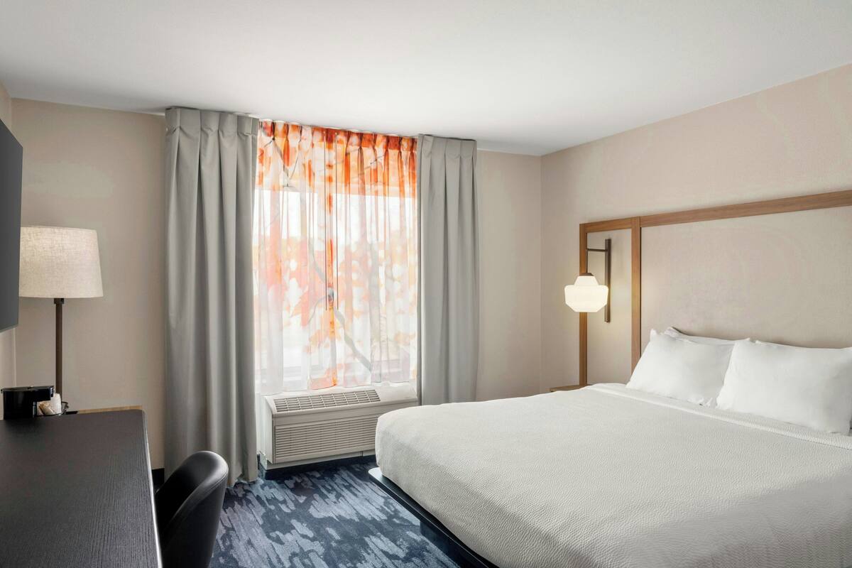 A hotel room with a large bed, a desk, a chair, a lamp, and a window with curtains.