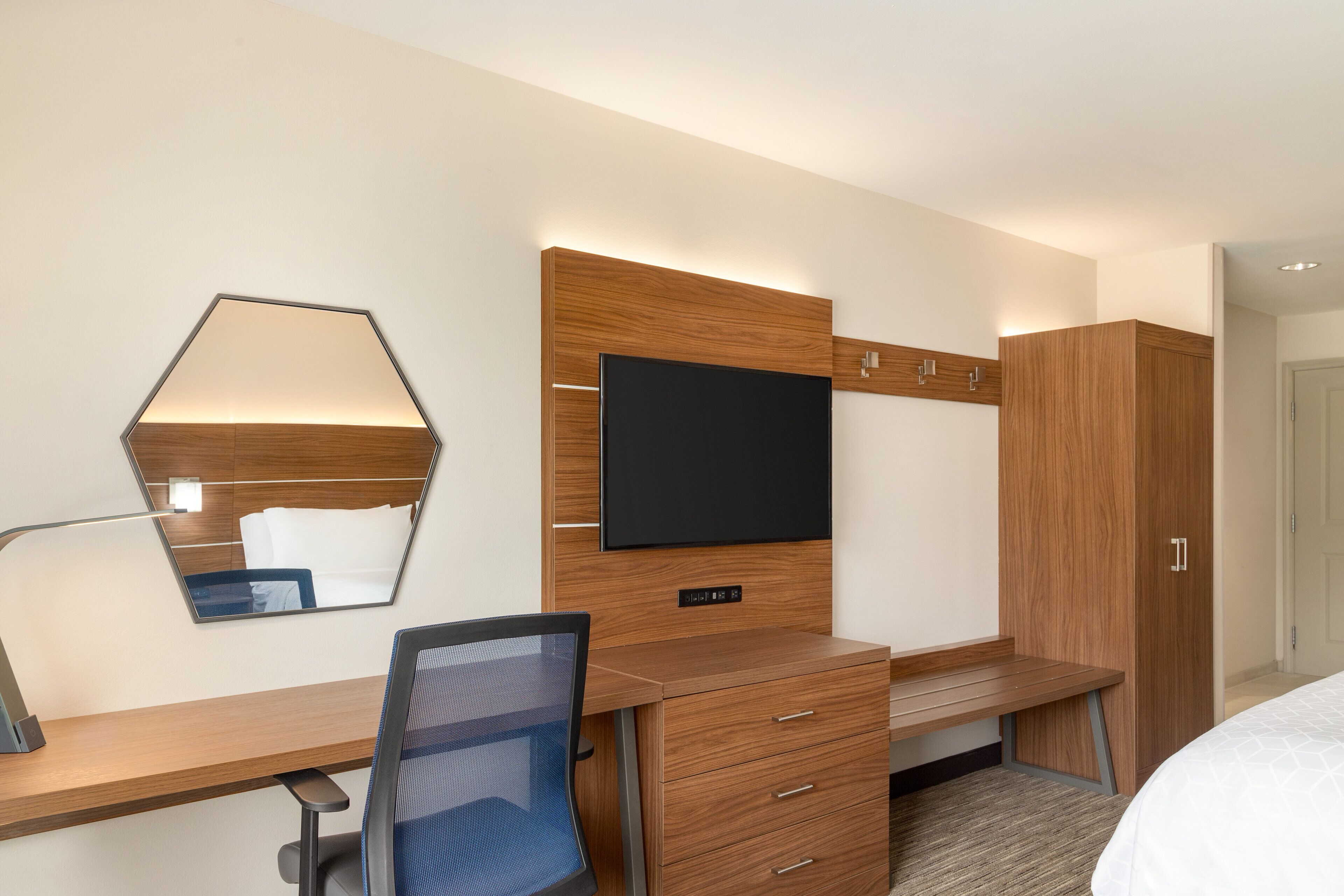A modern hotel room with a wooden desk, a flat-screen TV, a mirror, and a wardrobe.
