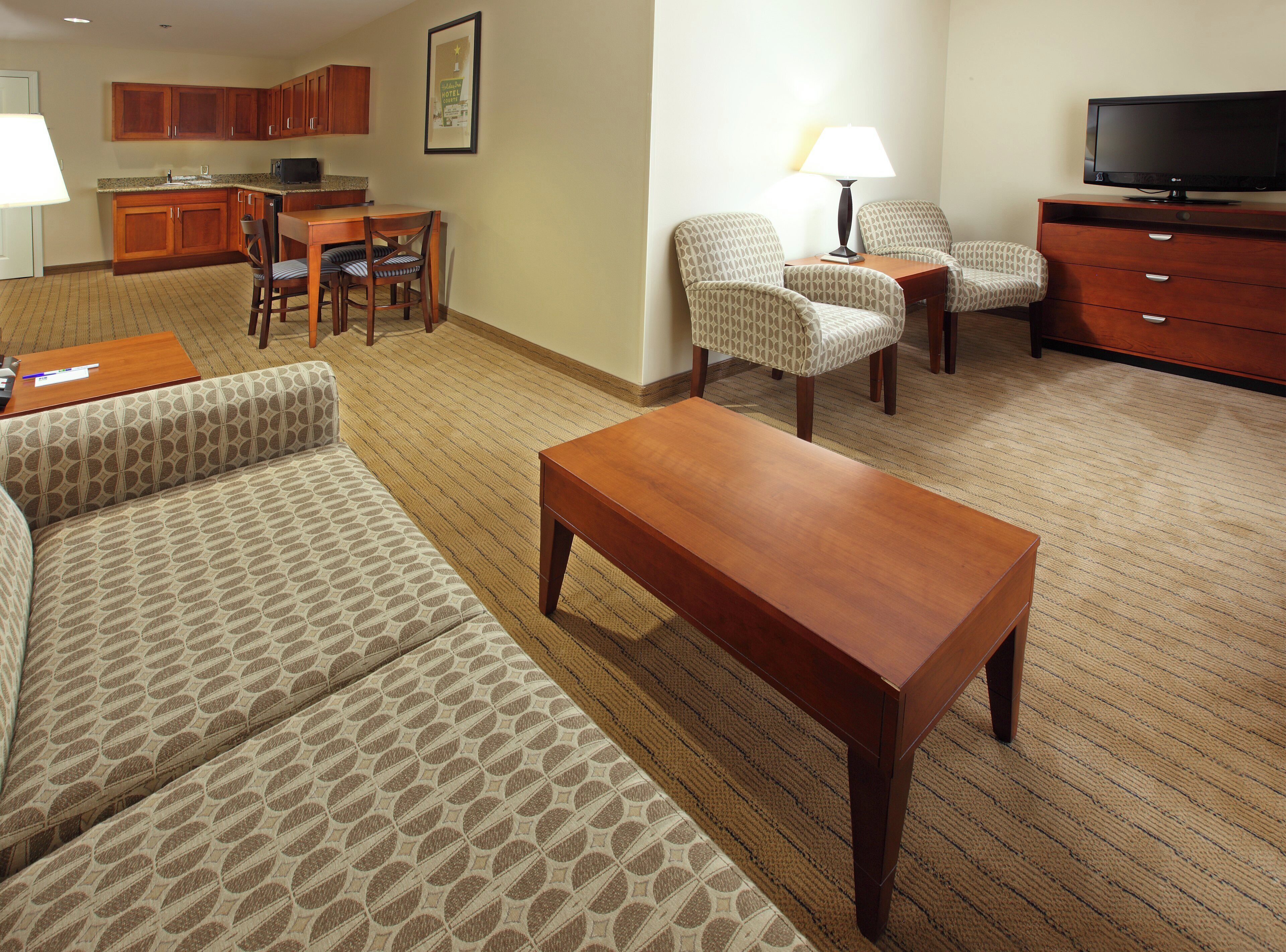 Habitación de hotel con sala de estar, una mesa de comedor, dos sofás estampados, una mesa de centro de madera, un televisor y una lámpara.