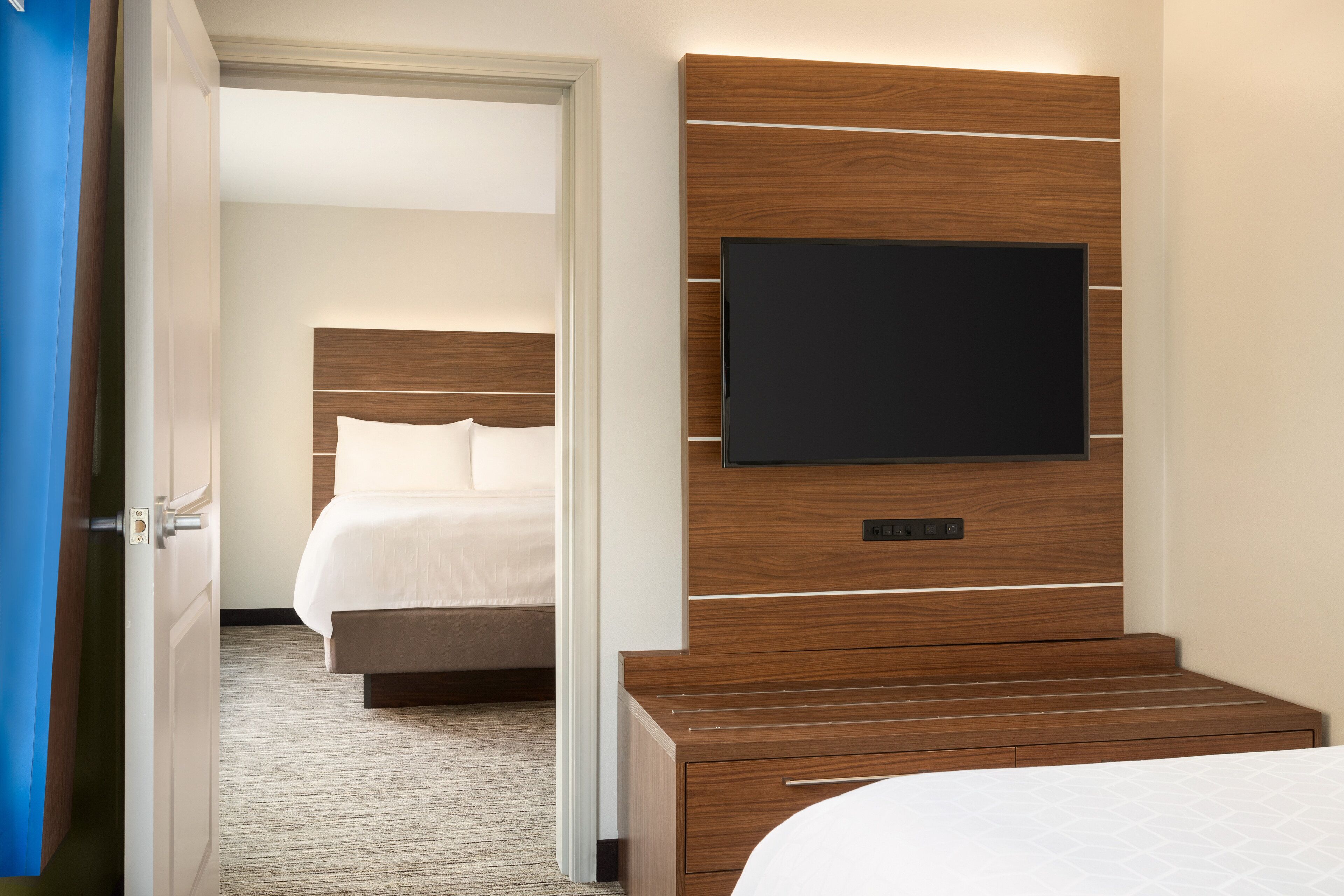 Habitación de hotel con cabecera de madera, un televisor de pantalla plana montado en la pared, ropa de cama blanca y suelo alfombrado.