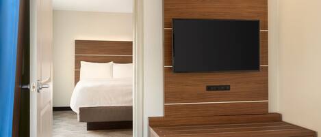 Habitación de hotel con cabecera de madera, un televisor de pantalla plana montado en la pared, ropa de cama blanca y suelo alfombrado.