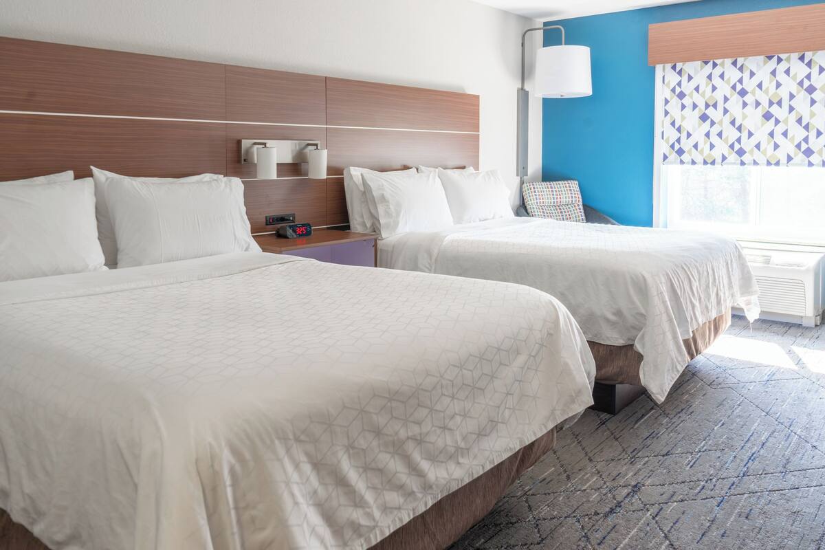 A hotel room with two beds, a nightstand, a lamp, a window with patterned curtains, and a blue accent wall.