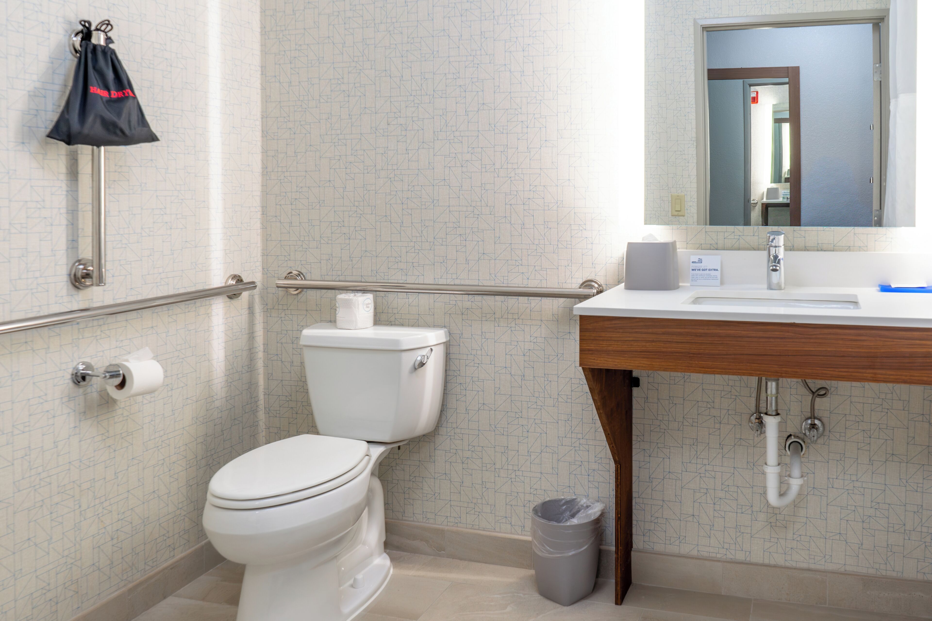 Un baño con inodoro, barras de apoyo, lavamanos, espejo y cubo de basura.