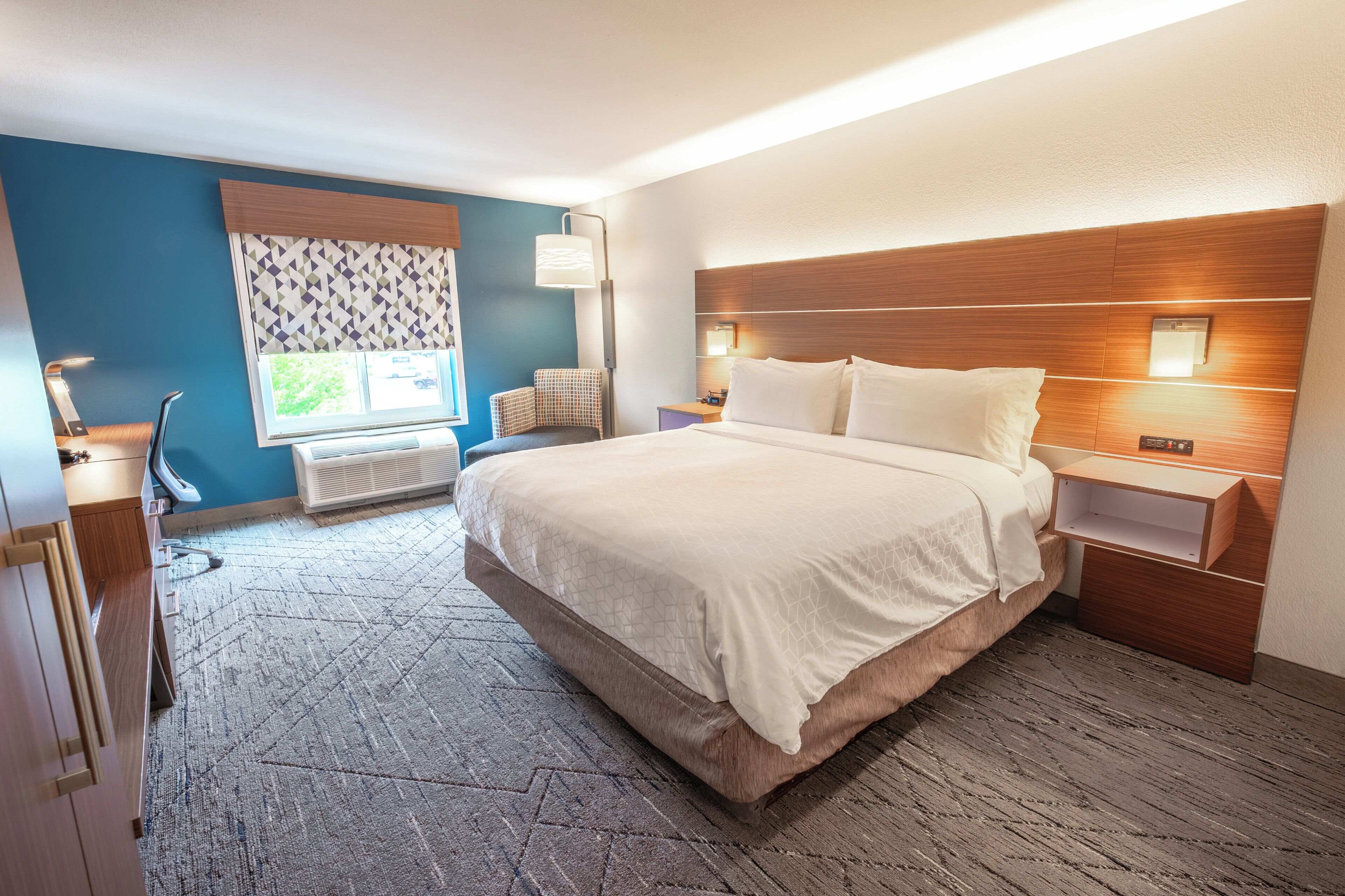 A hotel room with a large bed, a desk, a chair, a window with patterned curtains, and a wall-mounted lamp.