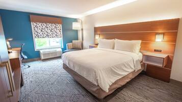 A hotel room with a large bed, a desk, a chair, a window with patterned curtains, and a wall-mounted lamp.