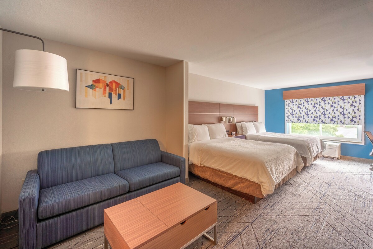 A hotel room with a sofa, a bed, a wooden table, a lamp, and a window with a view.