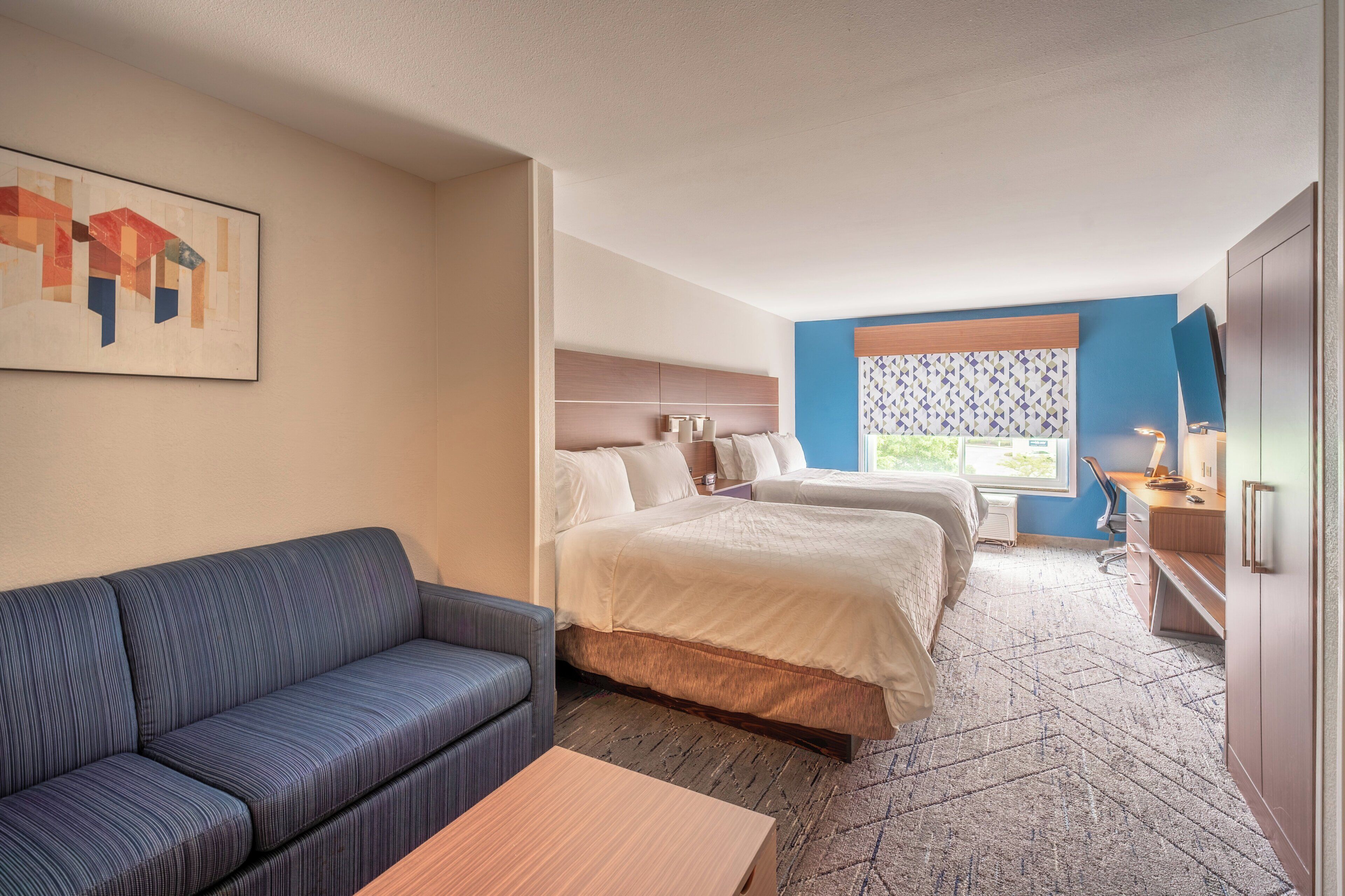 A hotel room with a sofa, two beds, a desk, and a window with patterned curtains.