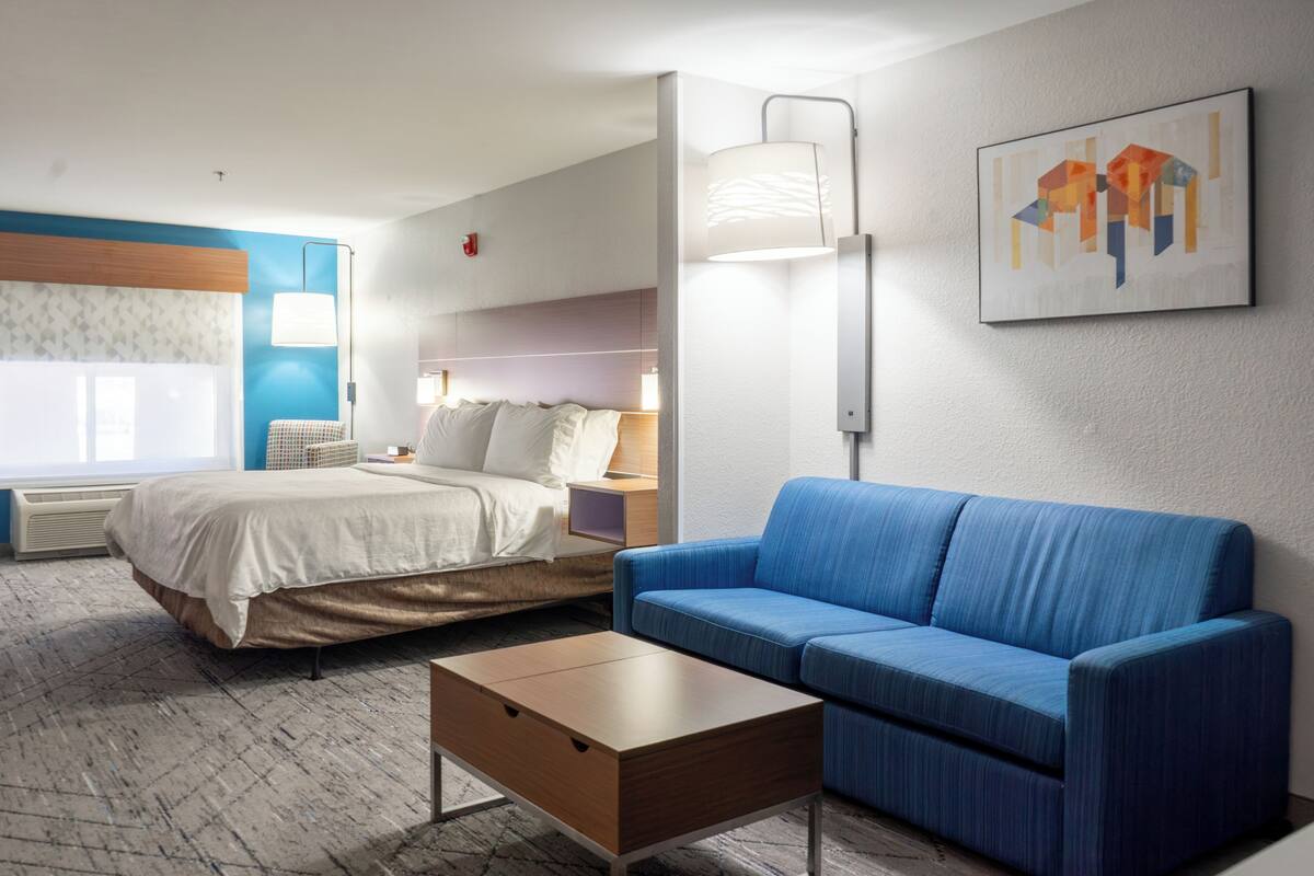 A hotel room with a bed, a blue sofa, a bedside table, a lamp, and a painting on the wall.