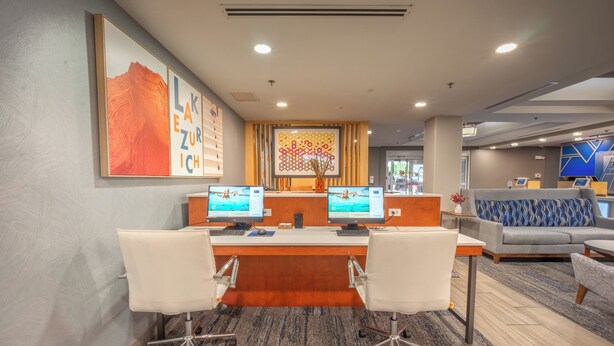 A reception desk with two computer monitors, a seating area with a sofa and armchairs, and a patterned carpet. - Holiday Inn Express Hotel & Suites, a Lake Zurich-Barrington by IHG (Lake Zurich)