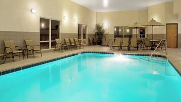 An indoor swimming pool with lounge chairs and umbrellas, surrounded by walls with windows and doors.
