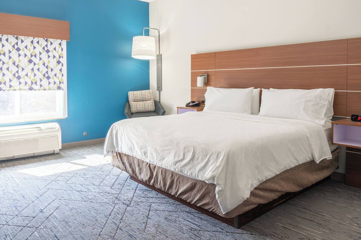 A hotel room with a bed, a nightstand, a chair, a lamp, and a window with patterned curtains.