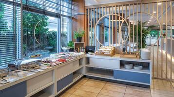 Ein reichhaltiges Buffet mit verschiedenen Speisen und einer Kaffeestation, umgeben von großen Fenstern und Zimmerpflanzen.