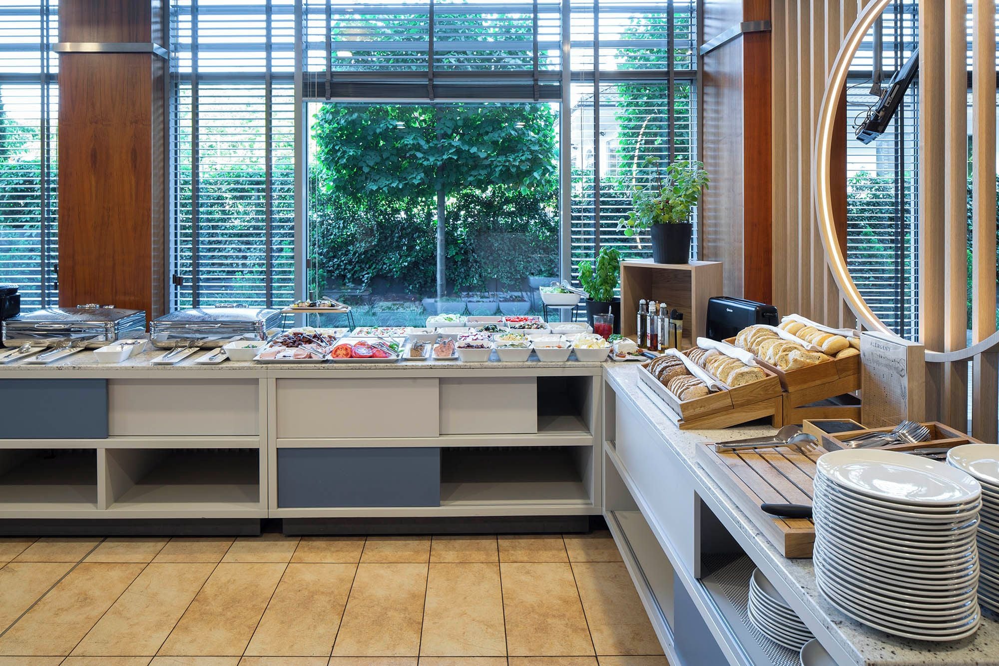 A buffet setup with various food items and utensils, a window with greenery view, and a tiled floor.