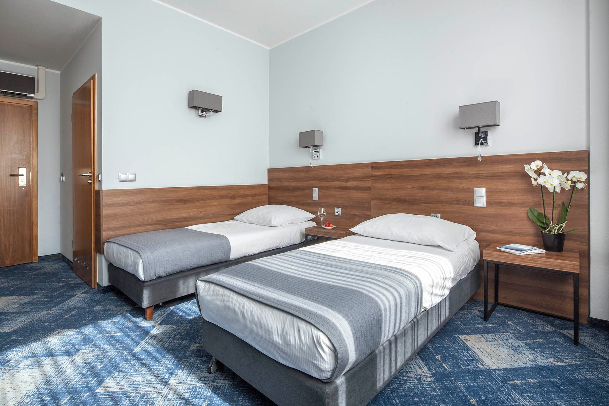 Une chambre d’hôtel avec deux lits, une table de chevet, un téléviseur fixé au mur et une orchidée en pot.