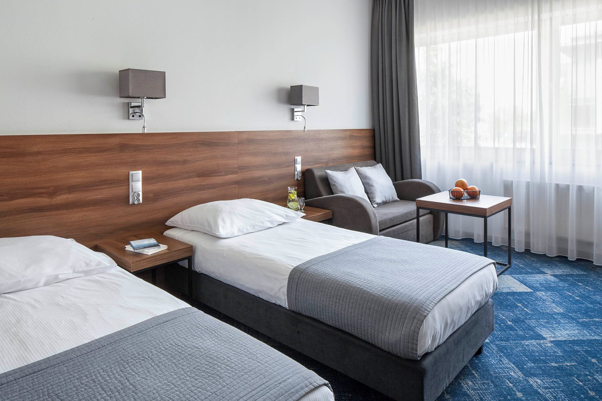 Quarto de hotel com duas camas, uma mesinha, um sofá e uma janela com cortinas.
