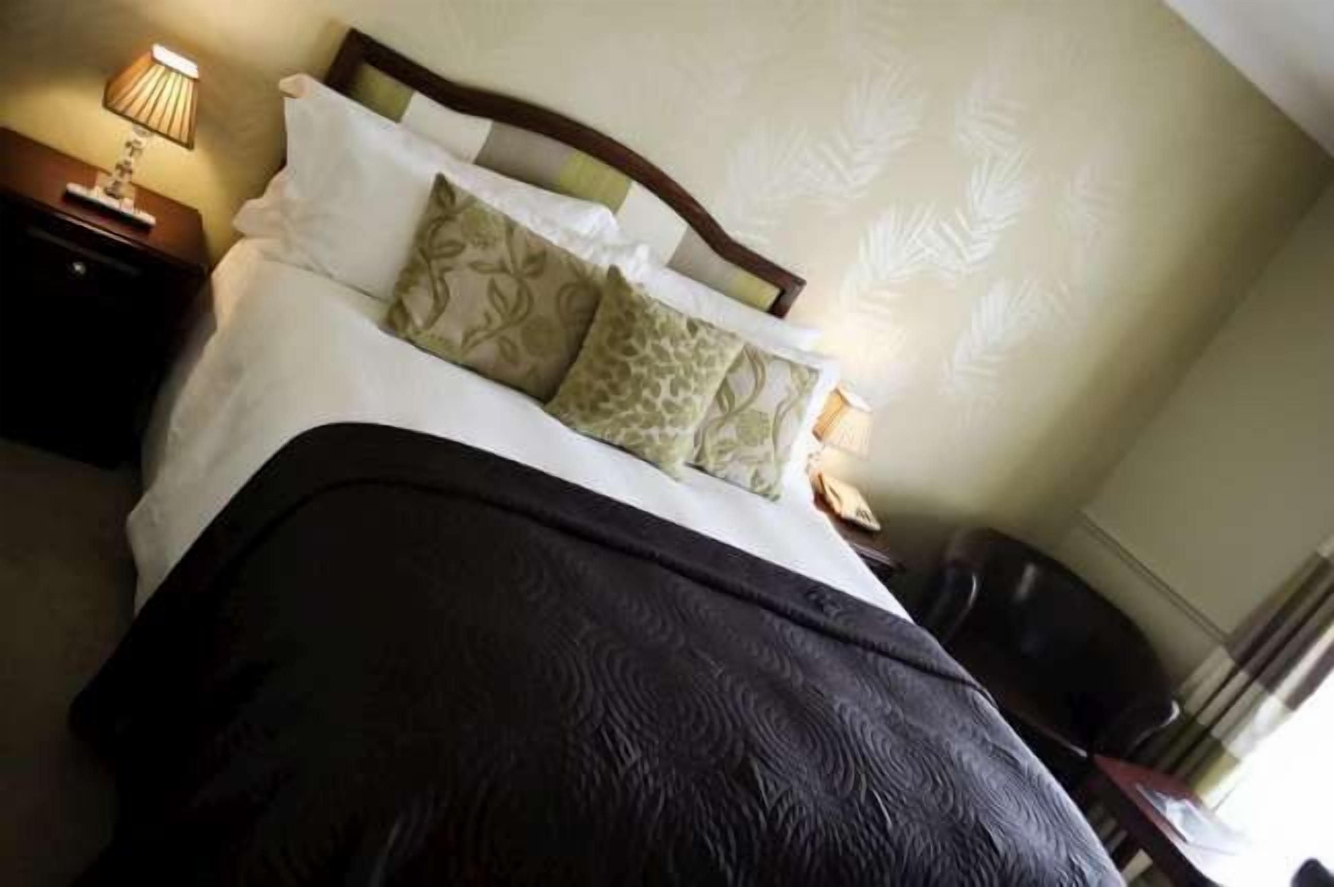 A neatly made bed with a dark patterned bedspread, a wooden headboard, and decorative pillows. There is a nightstand with a lamp and a small table beside the bed.