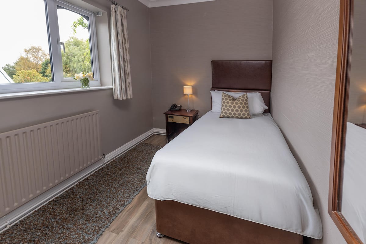 Ein Hotelzimmer mit einem Bett, einem Nachttisch, einer Lampe, einem Fenster mit Vorhängen und einem Heizkörper.