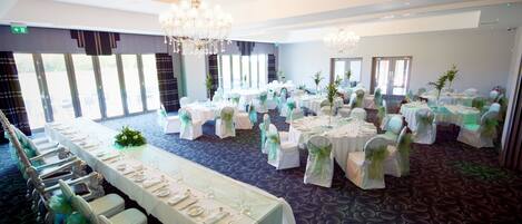 A banquet hall with long tables, chairs, chandeliers, and large windows.