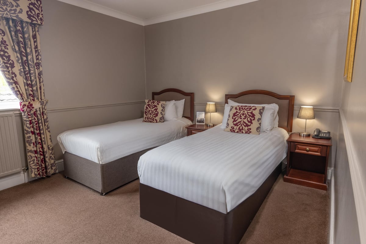 Ein Hotelzimmer mit zwei Einzelbetten, einem Nachttisch mit Lampe und einem Fenster mit Vorhängen.