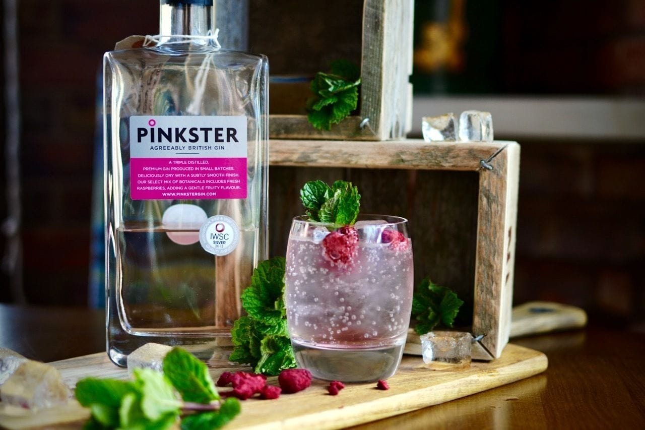 Une bouteille de gin Pinkster et un verre décoré d’une framboise, le tout posé sur une surface en bois.