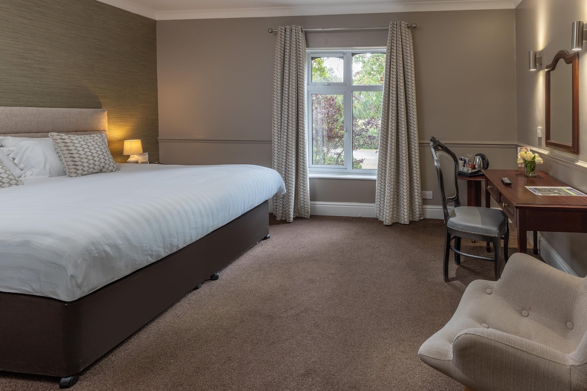 Ein Hotelzimmer mit einem großen Bett, einem Schreibtisch mit Stuhl, einem Fenster mit Vorhängen und einem Spiegel an der Wand.