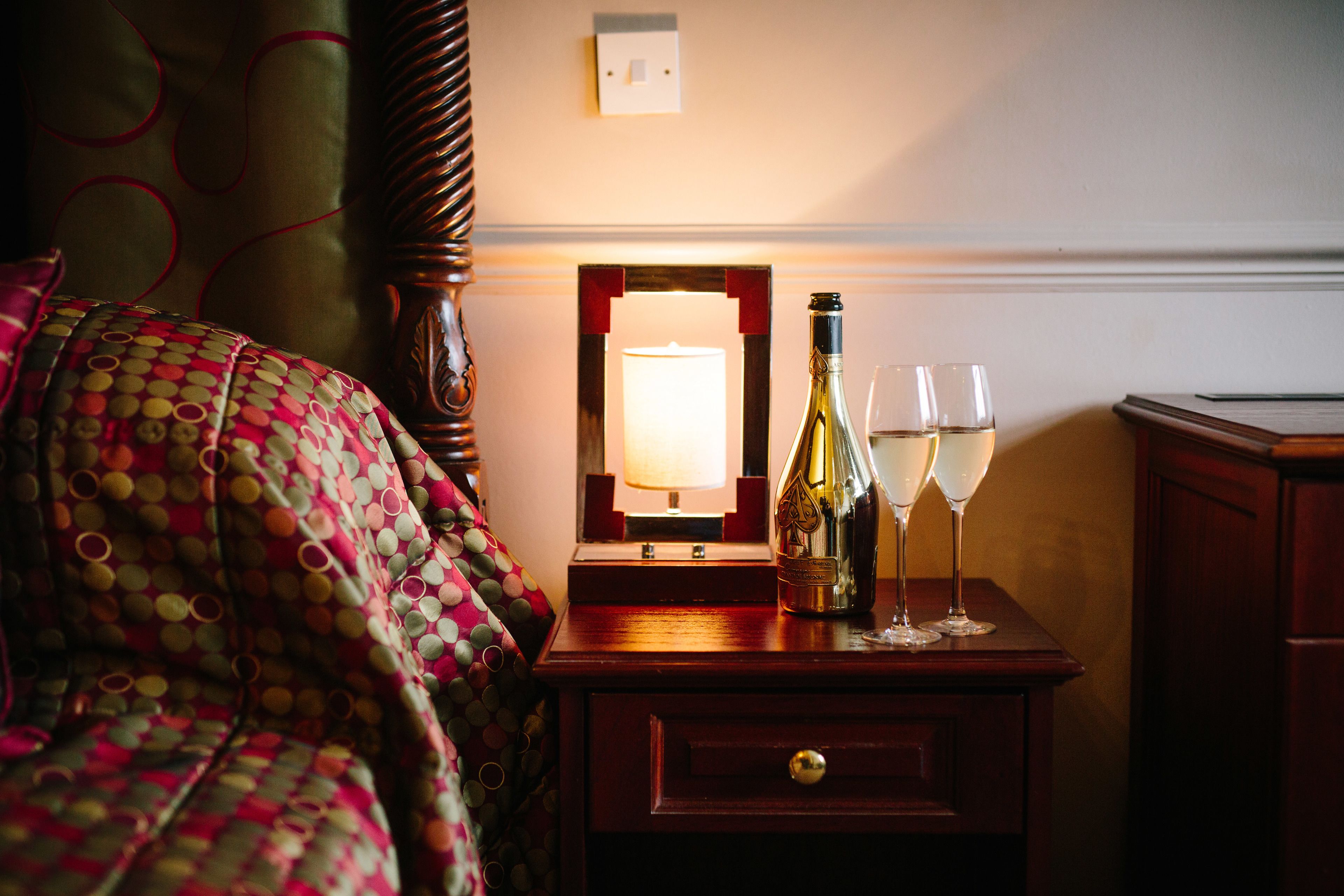 Un lit bien fait, recouvert d’une couverture à pois colorés, une table de chevet avec une bouteille de vin et deux verres, et un miroir avec un cadre rouge.