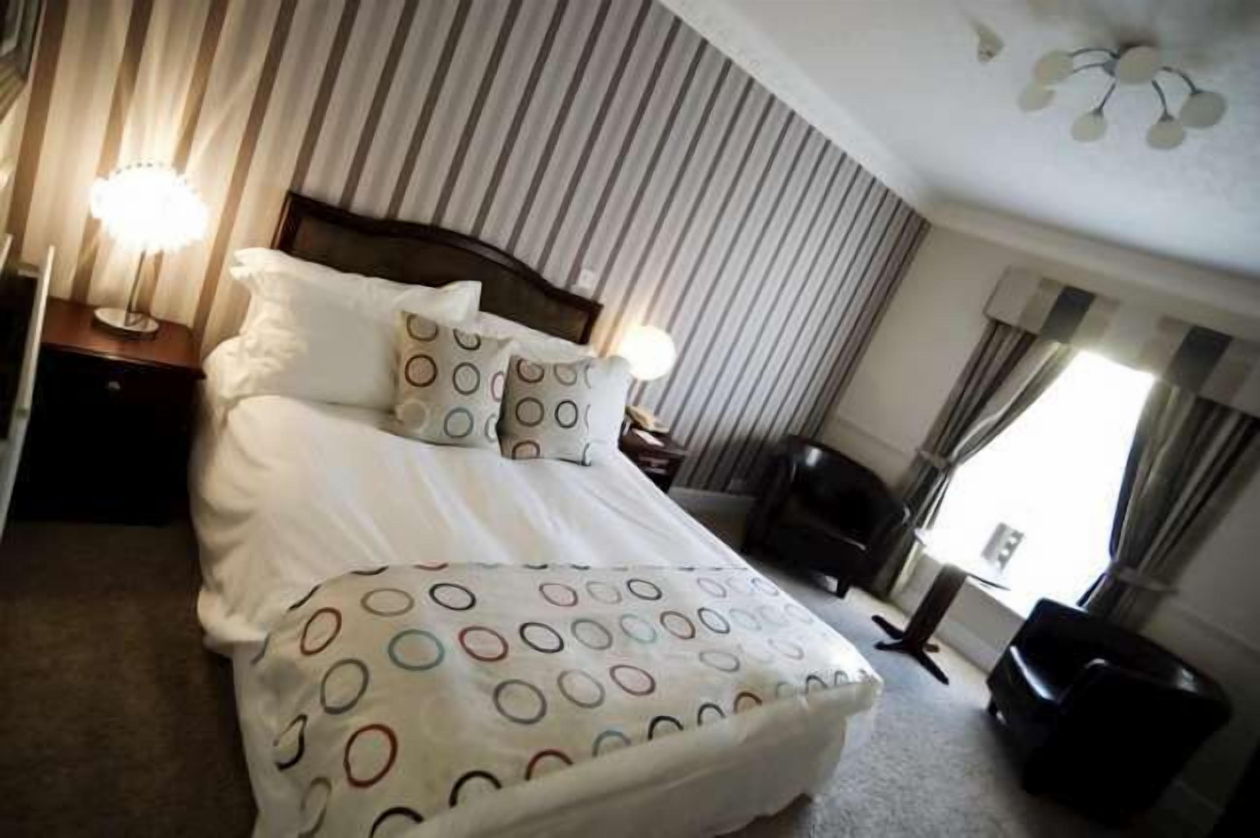 Een net opgemaakt bed met een beddengoed met patroon, een bedlampje en een stoel in een kamer met gestreepte gordijnen en een plafondlamp.