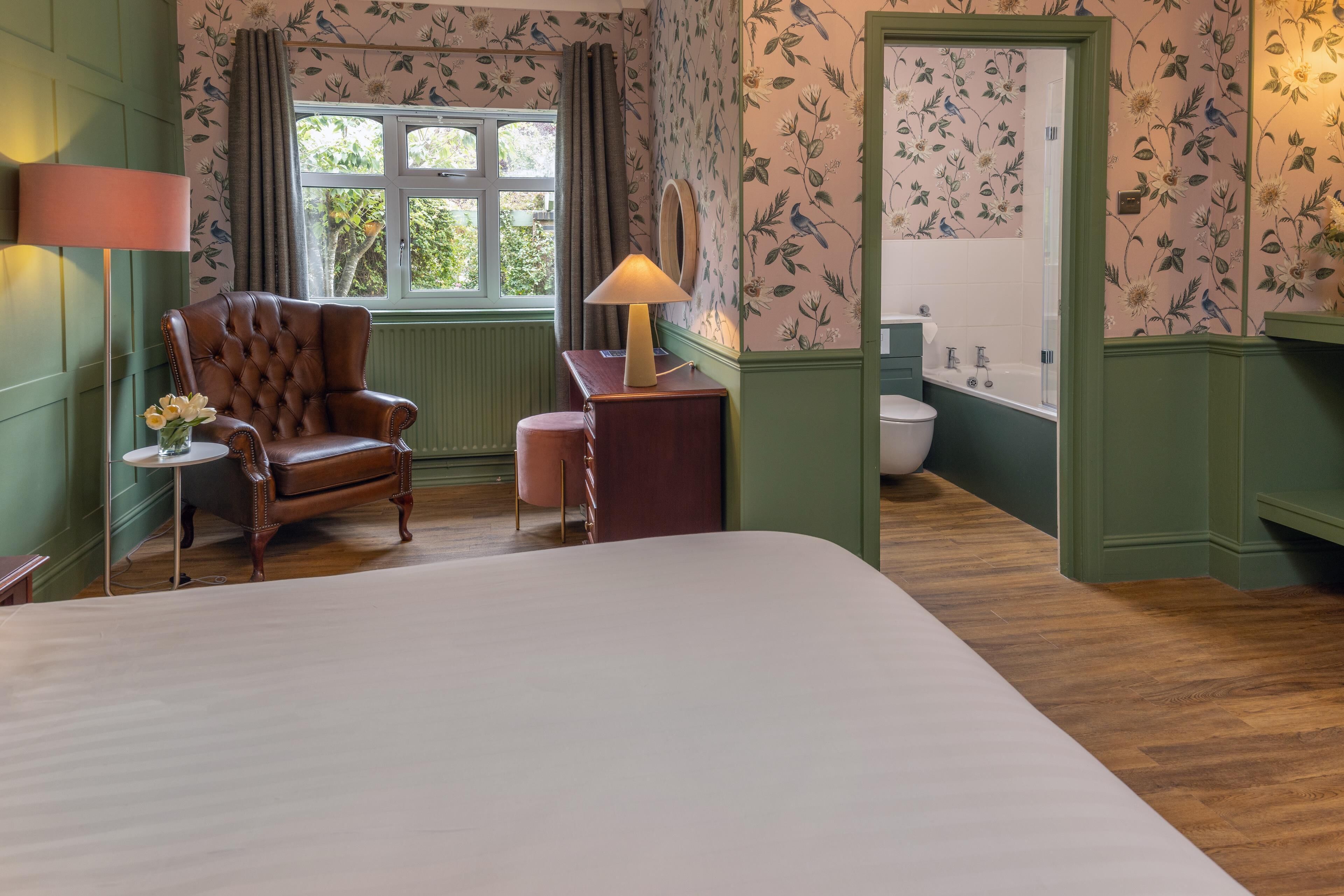 A bedroom with a large bed, a wooden chair, a small table with a lamp, and a view of the bathroom through an open door.