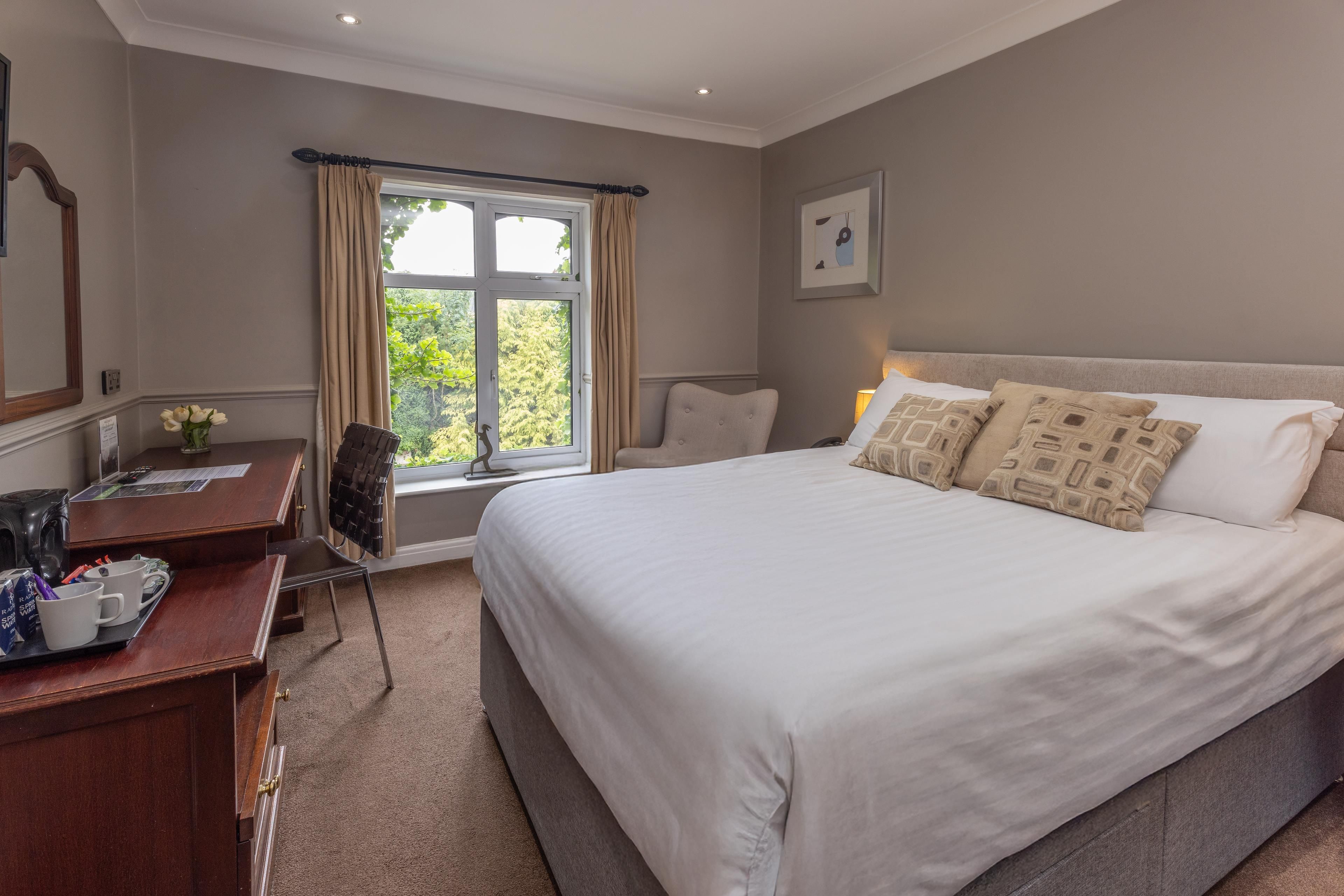 A hotel room with a large bed, a desk with a chair, a window with curtains, and a framed picture on the wall.