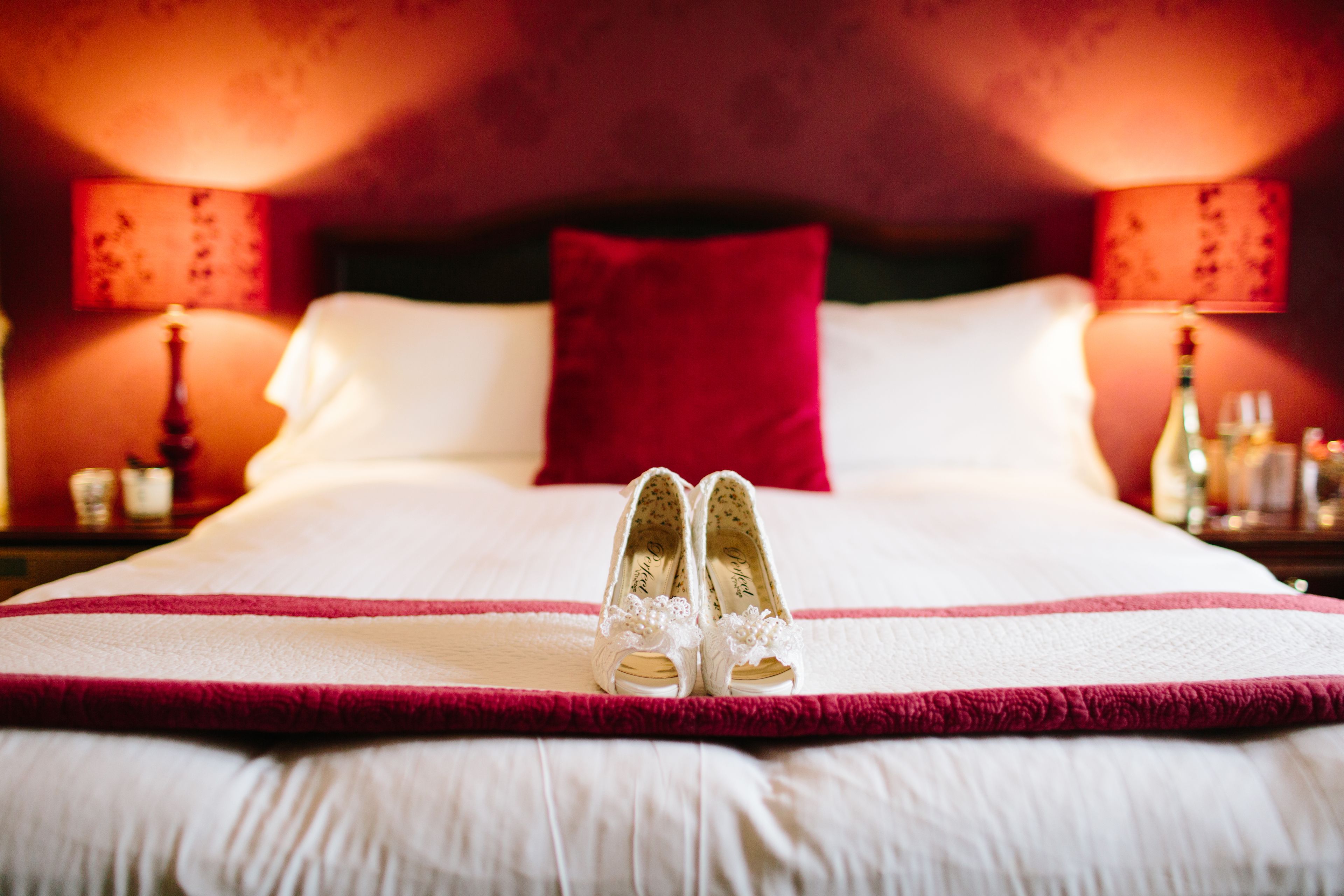 A bed with white linens and a red pillow, a pair of white shoes with floral details, and two bedside lamps with red shades.