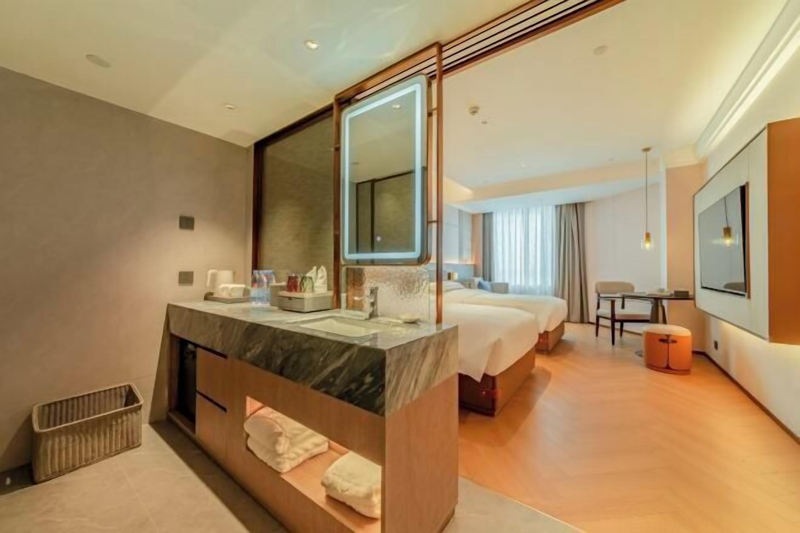 A modern hotel room with a large mirror, a marble vanity, a bed, a TV, and a seating area.