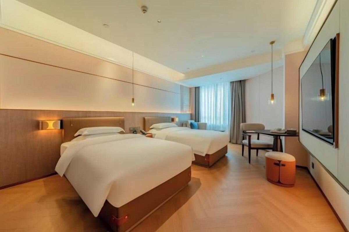 Ein modernes Hotelzimmer mit zwei Betten, einem Schreibtisch, einem Sessel und einem großen Fenster.