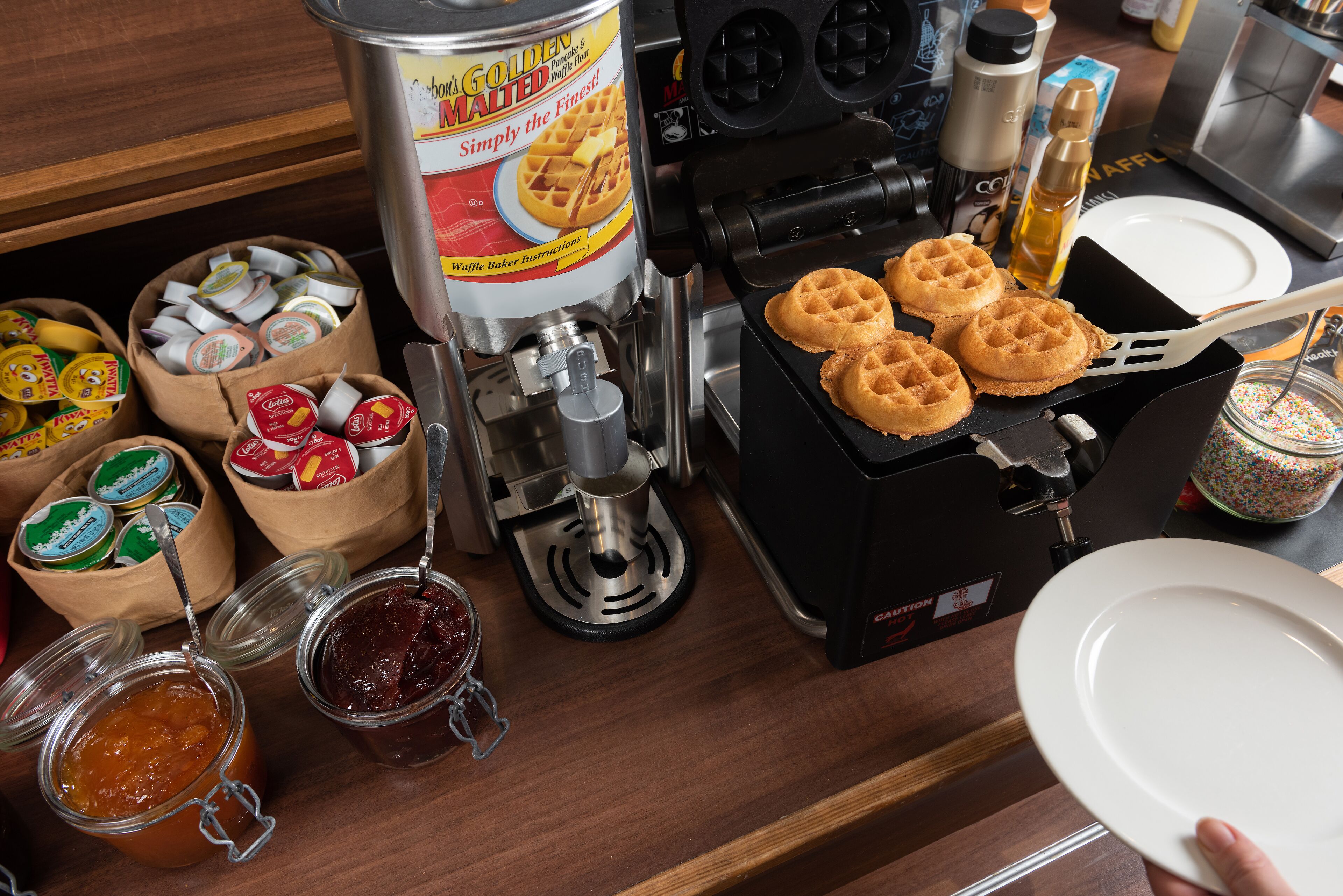 Een wafelijzer met wafels, een wafelbeslagdispenser en diverse toppings.