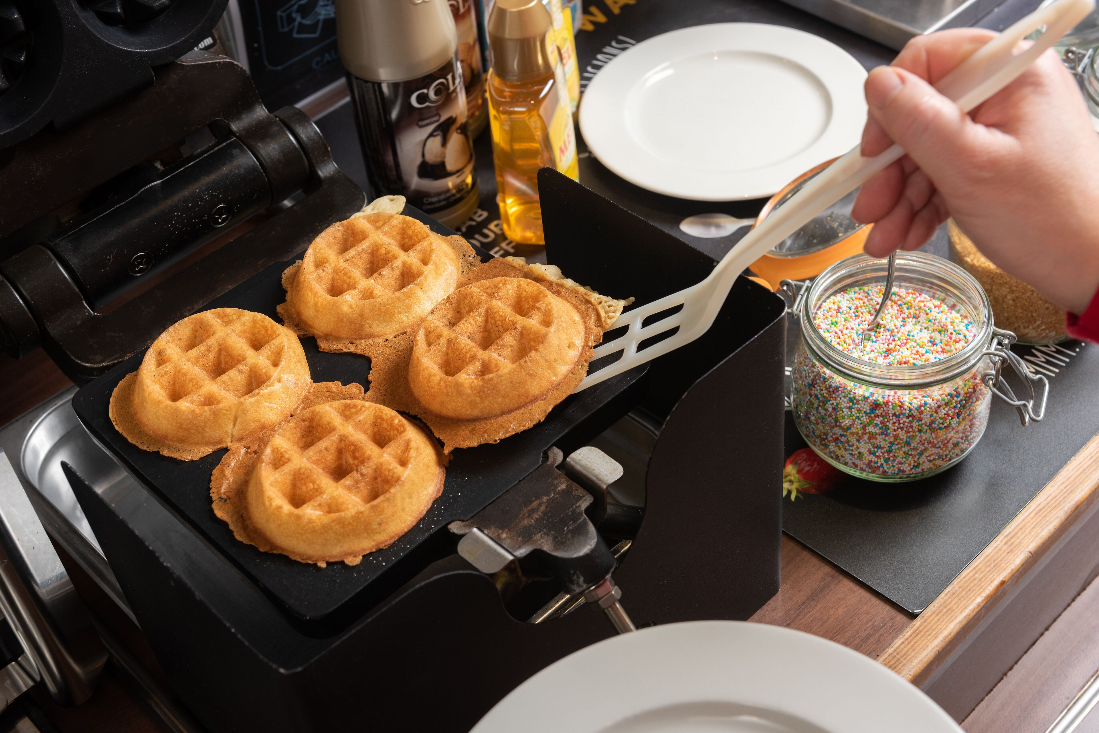 A waffle iron with waffles on a stove, a jar of sprinkles, and a white plate.
