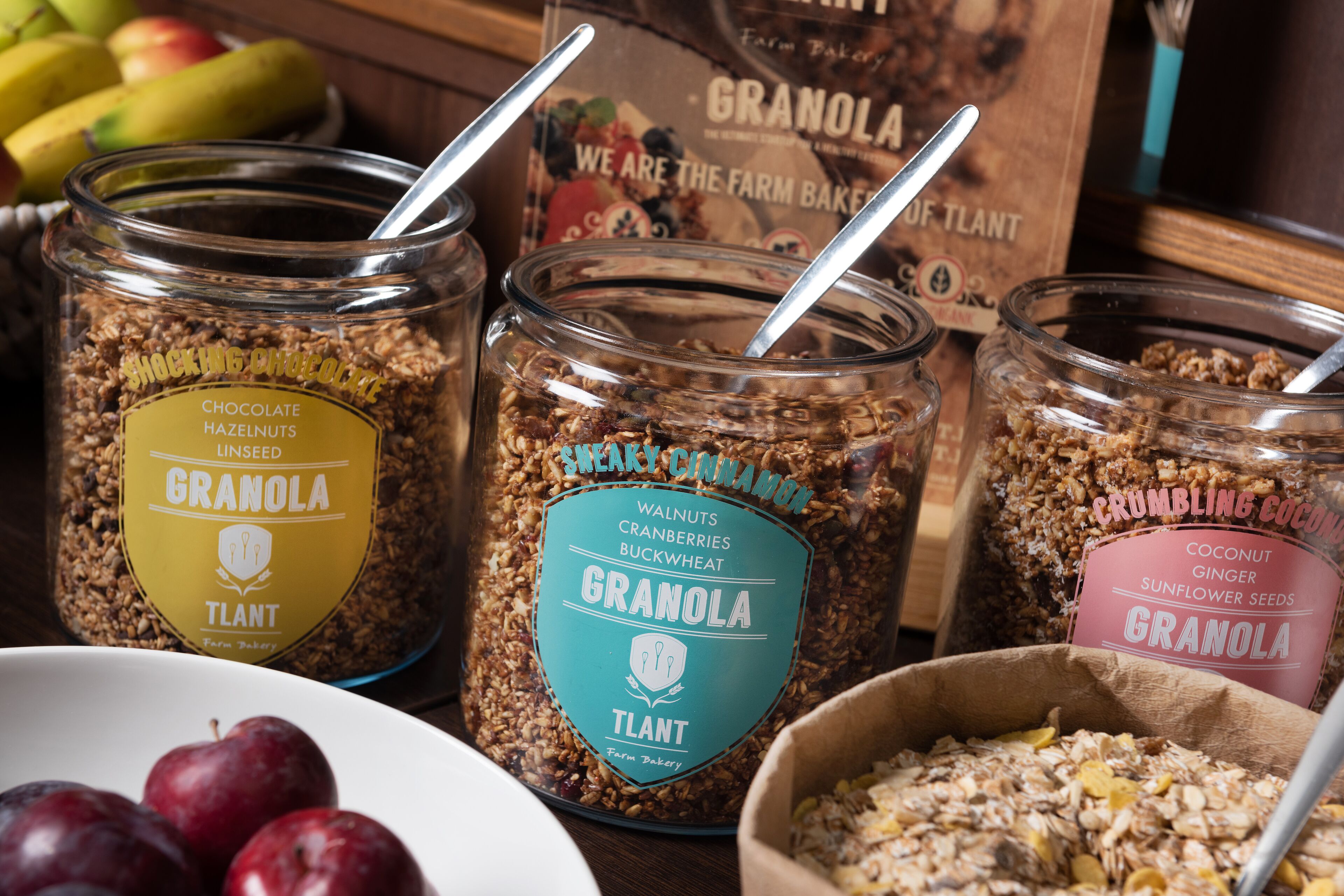 Three glass jars of granola with different flavors and ingredients, a bowl of plums, and a box of granola in the background.