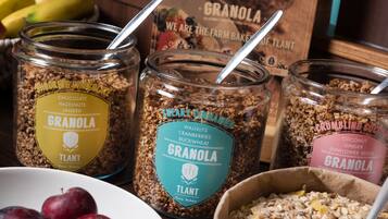 Three glass jars of granola with different flavors and ingredients, a bowl of plums, and a box of granola in the background.