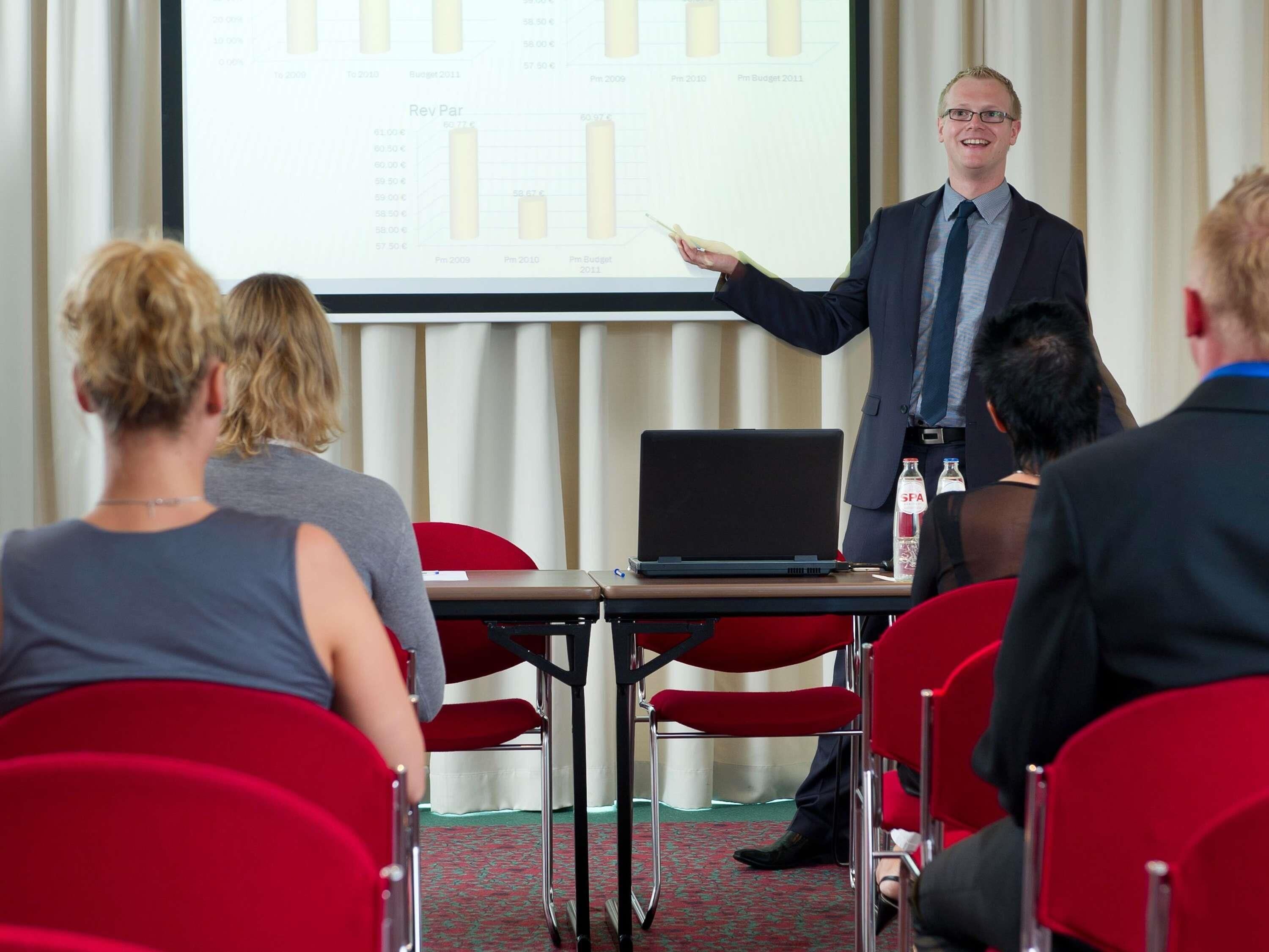 A conference room with a presentation screen, a laptop on a table, and attendees seated facing the presenter.