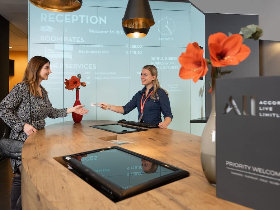 En receptionsdisk med en digital skærm, en træbordplade og en vase med blomster.