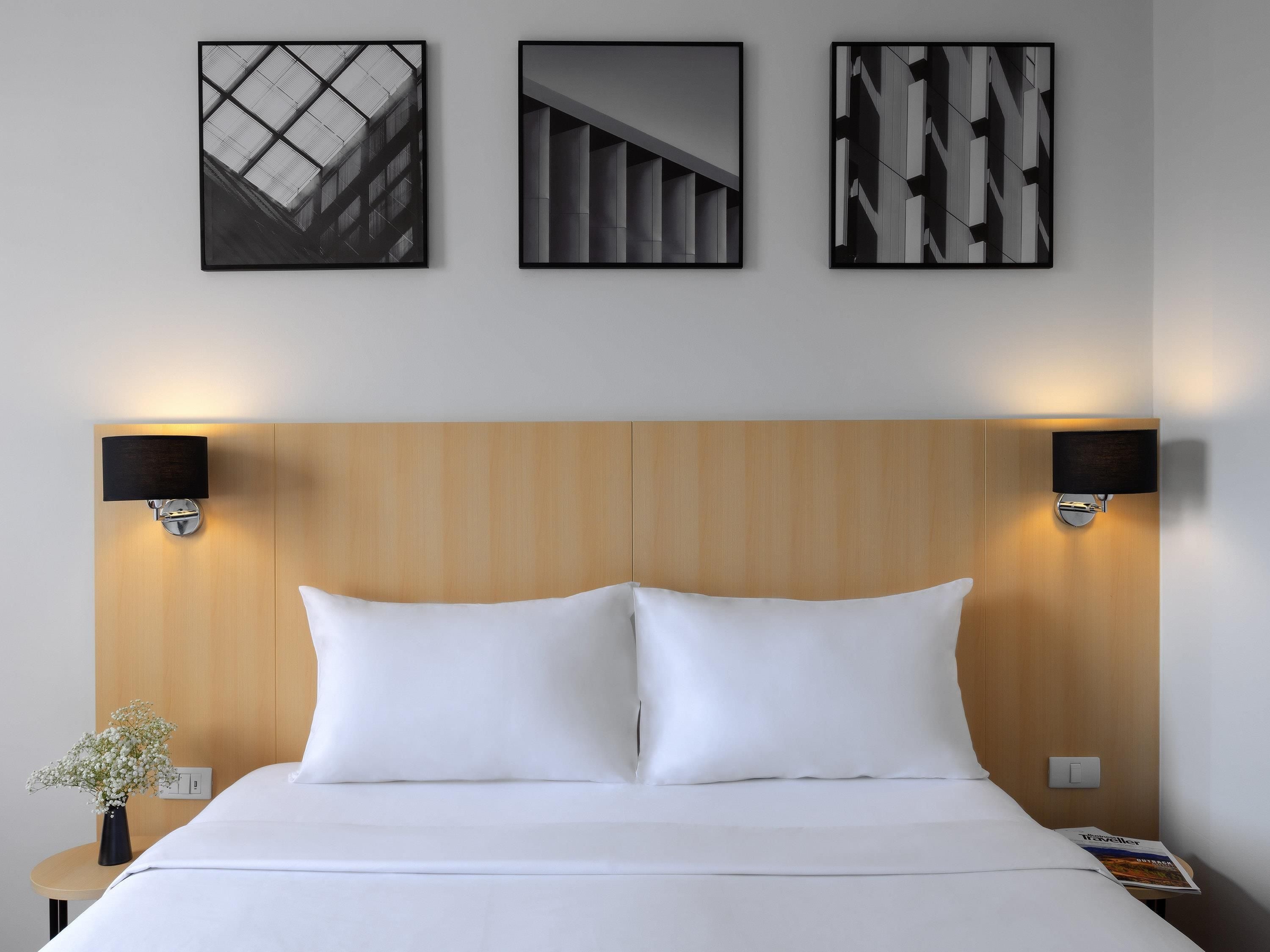 A hotel room with a wooden headboard, two wall-mounted lamps, a bed with white bedding, and three framed pictures on the wall.
