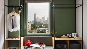 Een moderne kamer met een groot raam, een zithoek met een laptop, koptelefoon en een rode tas, en een bureau met een telefoon en waterkoker.