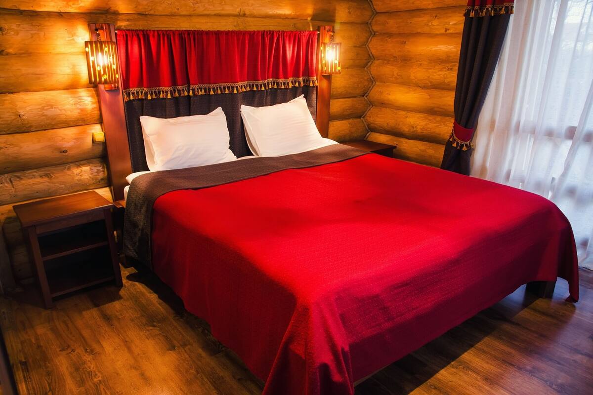A log cabin-style bedroom with a wooden headboard, a red bedspread, and a nightstand with a lamp.