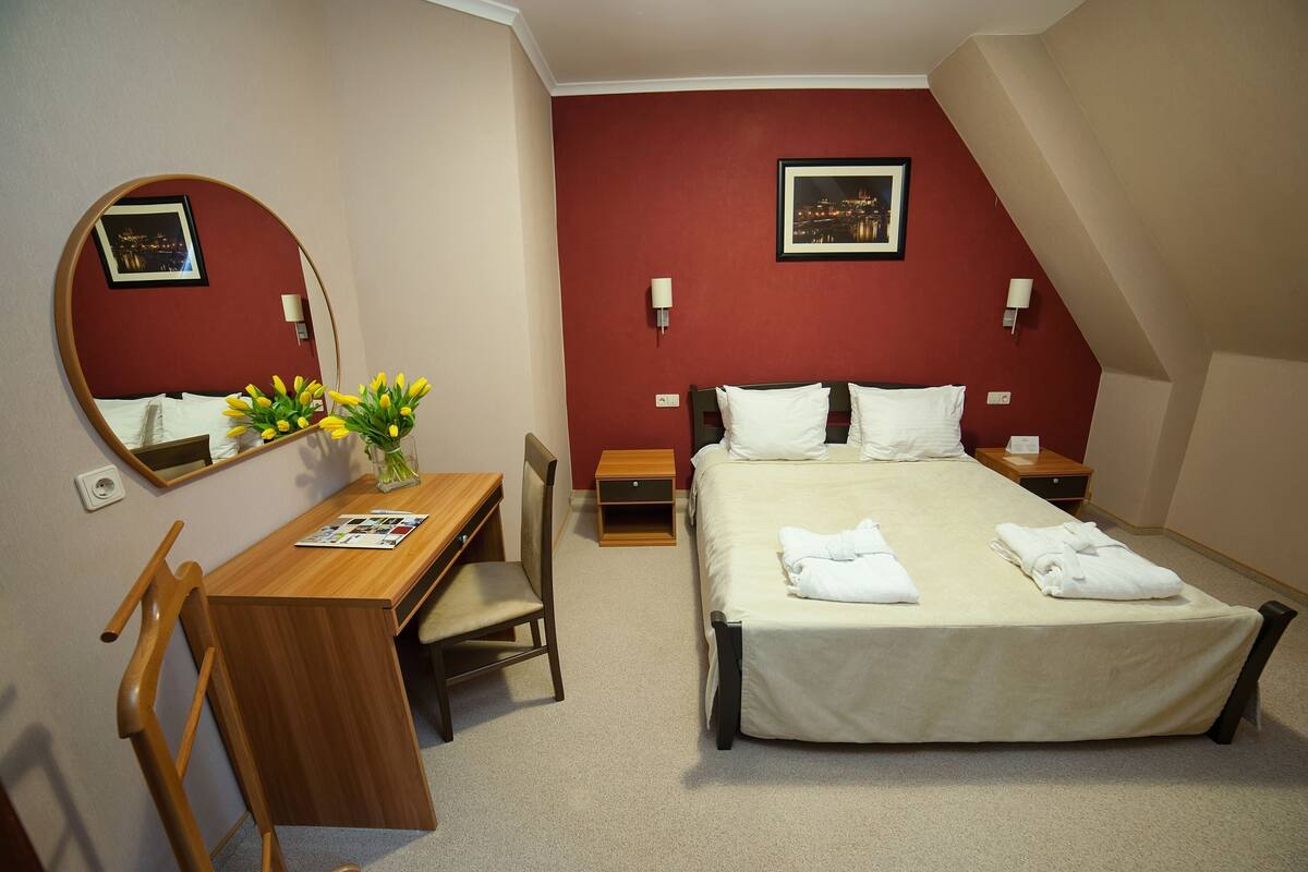 A hotel room with a bed, a desk, a chair, a mirror, and a vase with flowers.