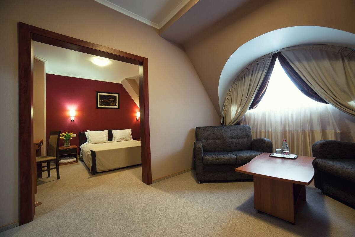 A hotel room with a bed, a desk, a chair, and a sofa. There is a window with curtains and a view of the outside.