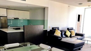Un espacio de sala moderno con una mesa de vidrio, un sofá de cuero negro con cojines estampados, y una zona de cocina con salpicadero verde.