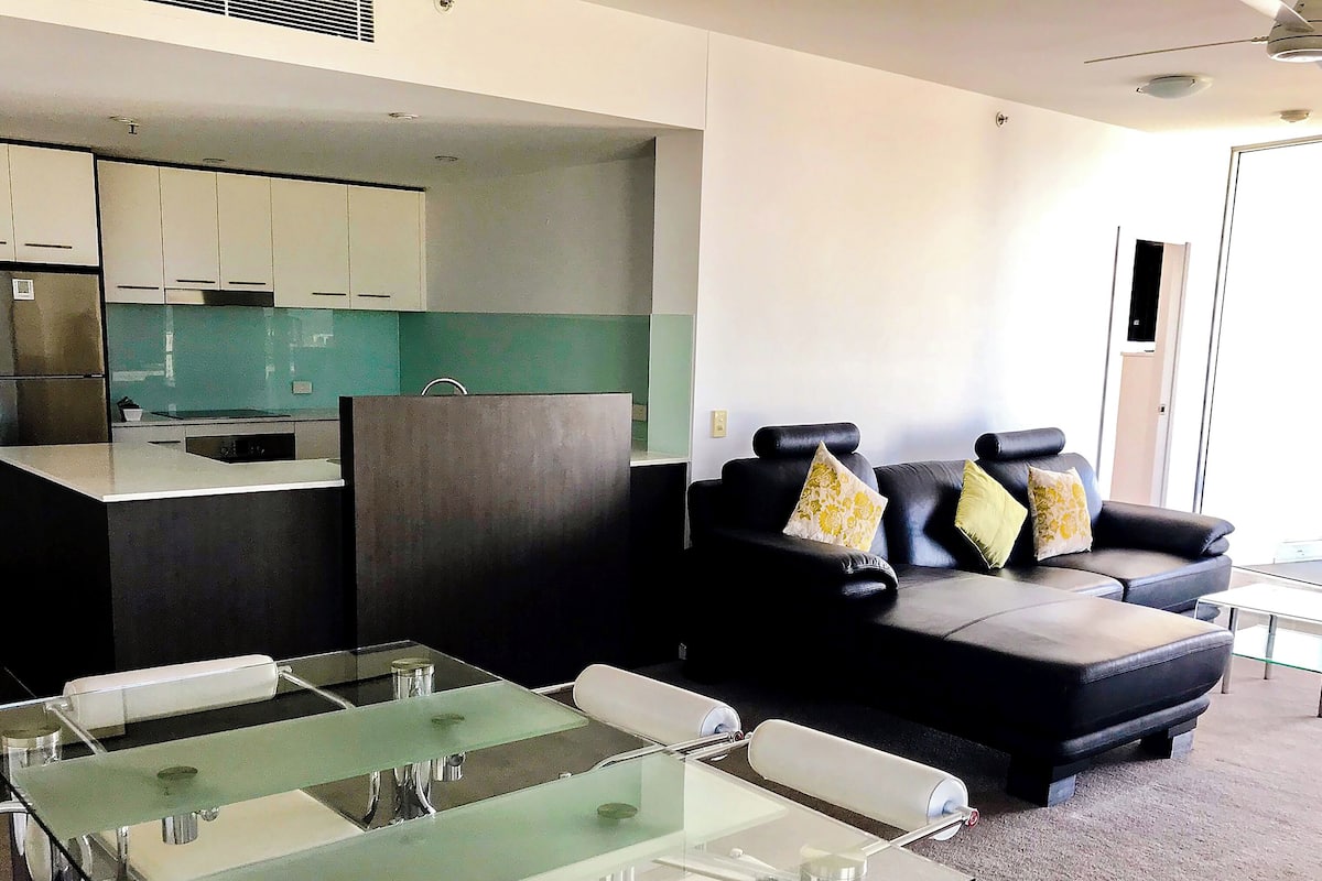 A modern living area with a glass-top table, a black leather sofa with floral cushions, and a kitchen area with a green backsplash.
